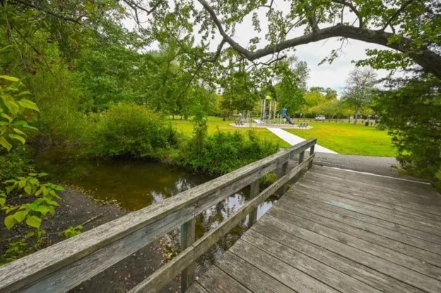 A park with a wooden pedestrian bridge over a waterway and a playground in the background.